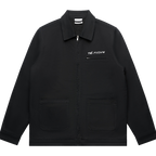 Front view of a black unisex relaxed fit jacket with a collar, front zip closure, and two lower front patch pockets. Features a white 'THE POISON' text print above a small zip chest pocket on the right side.